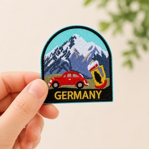 May include: An embroidered patch with an arched top featuring a red car, a yellow beer stein, and snow-capped mountains against a light blue sky. The word GERMANY is stitched in yellow capital letters at the bottom on a black background.
