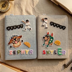 Könnte beinhalten: Zwei personalisierte Notizbuchhüllen. Eine ist hellblau mit einem Baseballspieler und einem Hundeaufnäher, dem Namen "ZEPHYR" in bunten Buchstaben und dem Wort "HAPPY". Die andere ist grau mit einem Hundeaufnäher, dem Namen "SKYLER" und dem Wort "HAPPY".