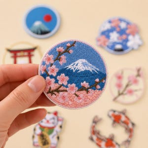 May include: A circular embroidered patch featuring a blue mountain with a snow-capped peak, framed by pink cherry blossom branches. The patch has a light pink border and is surrounded by other embroidered patches with various designs.