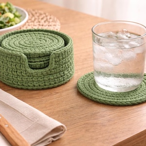 May include: A set of green crocheted coasters and a holder, with a glass of water on one coaster. The coasters are round and have a textured, spiral design. A fork and napkin are on the table.