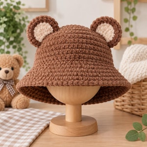 Op de afbeelding: Een bruine gehaakte bucket hat met twee ronde berenoren aan de bovenkant. De oren zijn gemaakt met een donkerbruine buitenring en een licht crèmekleurig midden. De hoed is gemaakt van zacht, getextureerd garen met een brede, afhangende rand.