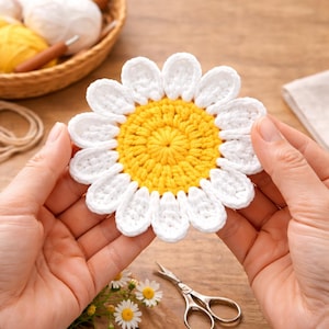 May include: A handmade crochet daisy with white petals and a yellow center. The flower is held between two hands, showcasing the detailed stitching. The craft project is set against a backdrop of craft supplies, including yarn and scissors.