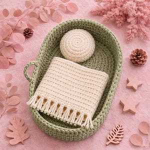 May include: An oval-shaped sage green crocheted basket containing a cream-colored crocheted blanket with fringe trim and a small, round, cream-colored crocheted pillow. The basket features a single side handle and a thick, textured weave.