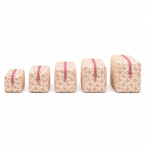 Peut inclure: Cinq trousses rectangulaires à fermeture éclair disposées par taille croissante. Chaque trousse présente une base en tissu crème clair avec un motif floral dense et répétitif dans des tons rose, rouge et vert, avec une fermeture éclair rose.