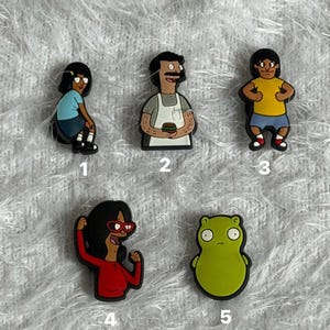 May include: Five colourful shoe charms featuring cartoon characters from Bob's Burgers. The set includes a girl in a blue shirt, a man with a moustache in an apron holding a burger, a boy in a yellow shirt, a girl in a red shirt with glasses, and a green creature.