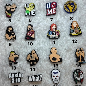 May include: A collection of twenty-one colorful PVC shoe charms featuring various WWE wrestling logos, championship belts, and character illustrations. The charms include text such as "U CAN'T SEE ME", "Austin 3:16", and "What?" arranged in a grid pattern.