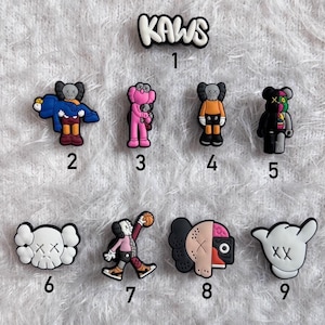 May include: A selection of KAWS-themed shoe charms in various colours and designs. The charms feature the KAWS logo and cartoon figures with signature X-shaped eyes. The figures are in pink, black, orange, and white.