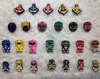 May include: A collection of colorful Power Ranger-themed shoe charms. The charms feature various characters in different colors, including red, blue, yellow, pink, green, black, and orange. Some charms are shaped like the Ranger's helmets.