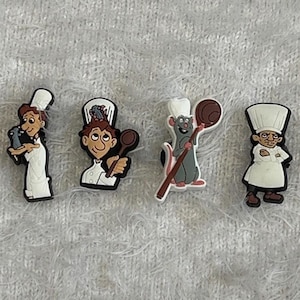 May include: A set of four shoe charms featuring cartoon characters in chef attire. The designs include a person holding a small grey creature, a person holding a wooden spoon, a grey rat holding a spoon, and a stern-looking chef with arms crossed.