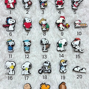 May include: A collection of twenty-three colourful shoe charms featuring Snoopy and Woodstock in various outfits and activities, numbered one through twenty-three. Each charm is a small, detailed figure made of rubberised material with a flat back.