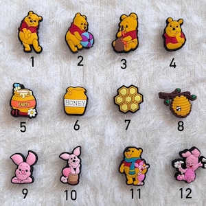 Op de afbeelding: Een verzameling kleurrijke schoenbedels met personages en symbolen uit een populaire kinderfranchise. De bedels bevatten een gele beer, een roze varken, een honingpot en een bord met de tekst "100 Acre Wood."