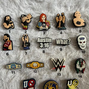 May include: A collection of black, white, red, and gold WWE-themed charms. The charms feature wrestler likenesses, championship belts, and the WWE logo. Some charms have text that reads "Austin 3:16", "What?", "U ME", and "Hustle Loyalty Respect".