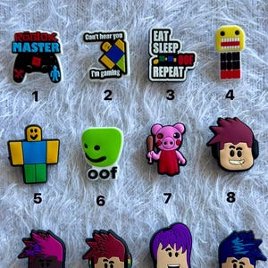 May include: A set of twelve colourful shoe charms featuring various gaming-themed designs, including characters, text like Roblox Master, Can't hear you I'm gaming, Eat Sleep Repeat, and Oof, numbered one through twelve, arranged in three rows of four.