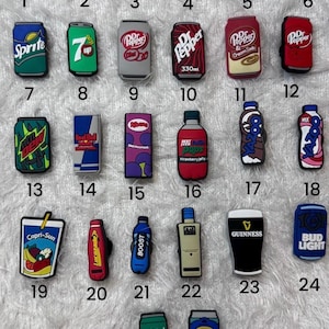 May include: A collection of twenty-six colorful rubber shoe charms shaped like various beverage cans, bottles, and cartons, each numbered from 1 to 26. The designs feature logos for brands including Coca-Cola, Pepsi, Fanta, Sprite, Dr. Pepper, and Guinness.