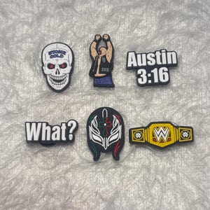 Könnte beinhalten: Eine Sammlung von sechs bunten Schuhanhängern. Die Anhänger zeigen einen Totenkopf mit dem Text "STONE COLD", eine Wrestlerfigur, den Text "Austin 3:16", den Text "What?", eine Wrestling-Maske und einen Meisterschaftsgürtel mit dem "W"-Logo.