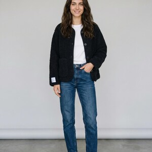 Könnte beinhalten: Eine Person trägt eine schwarze, strukturierte Jacke über einem weißen T-Shirt und einer blauen Jeans. Die Jacke hat eine Knopfleiste und zwei Taschen. Die Jeans ist ein klassischer, gerader Schnitt.