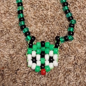 May include: A beaded necklace featuring a pendant with a pixelated design. The necklace is made of green and black beads, with a pendant in green, white, black, and red. The necklace is displayed on a brown surface.
