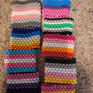 May include: A collection of colorful beaded bracelets in various pride flag color schemes. The bracelets are made of small, round beads strung together, showcasing a range of colors including pink, blue, purple, black, white, and yellow. The bracelets are displayed on a neutral-toned surface.