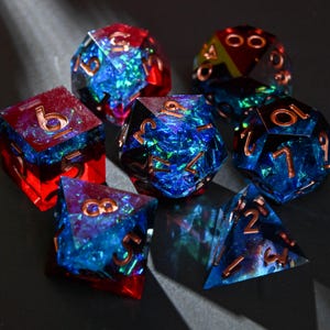 Op de afbeelding: Een set van zeven polyhedrale dobbelstenen in verschillende vormen, waaronder een d4, d6, d8, d10, d12, d20 en een percentieldobbelsteen. De dobbelstenen zijn gemaakt van heldere hars met blauwe, rode en groene iriserende insluitsels en koperkleurige cijfers.