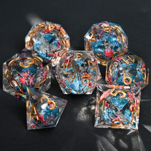 Könnte beinhalten: Set aus sieben transparenten, polyedrischen Würfeln für Tabletop-Spiele. Die Würfel haben einen blauen Glitzerkern, goldene Zahlen und rote und schwarze florale Akzente. Das Set enthält einen D4, D6, D8, D10, D12, D20 und einen Prozentwürfel.