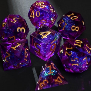 May include: Set of seven polyhedral dice in shades of purple and blue with gold numbering. The dice are made of a translucent material, with a glittery, galaxy-like effect. The dice are used for tabletop role-playing games.