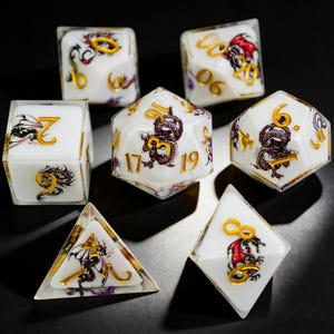 Puede incluir: Juego de siete dados poliédricos blancos con numeración dorada e ilustraciones de dragones. Los dados son para juegos de mesa y tienen un acabado de resina transparente. El arte del dragón es negro y rojo.