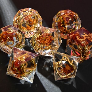 May include: Set of seven clear resin dice with gold-colored numbers and internal accents. The dice are various polyhedral shapes, including a d4, d6, d8, d10, d12, d20, and a percentile die. The dice have a sparkling, gem-like appearance.