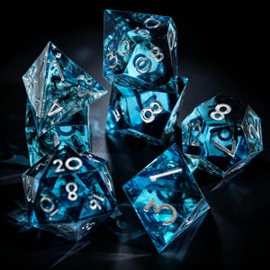 Puede incluir: Juego de siete dados poliédricos azules con números plateados. Los dados transparentes tienen un diseño interno ahumado y arremolinado. Los dados están colocados sobre una superficie oscura, para juegos de rol.