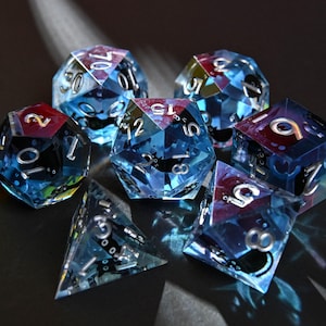 Könnte beinhalten: Ein Satz von sieben Würfeln in verschiedenen Formen, darunter ein d4, d6, d8, d10, d12, d20 und ein Prozentwürfel. Die Würfel sind transparent mit einem blau-schwarzen Farbschema und silbernen Zahlen. Die Würfel haben ein Katzendesign im Inneren.