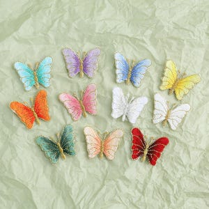Puede incluir: Un conjunto de parches bordados con forma de mariposa en varios colores, incluyendo turquesa, morado, azul, amarillo, naranja, rosa, blanco, verde, melocotón y rojo. Cada mariposa tiene un borde dorado.