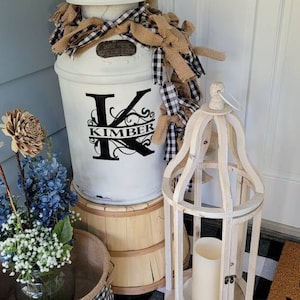 May include: A white milk can with a black and white checkered ribbon and a black vinyl decal with the letter K and the name Kimber. The can is sitting on a wooden barrel. A white wooden birdcage lantern is on the right. A black and white checkered rug is on the floor.