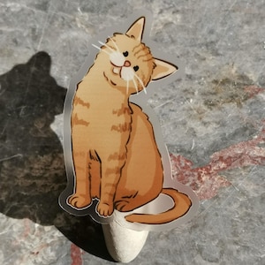 Op de afbeelding: Een gestanste sticker met een cartoonillustratie van een oranje cyperse kat met donkere strepen, witte snorharen en een schuin gehouden kop. De kat zit in een zittende houding met de staart om het lichaam gekruld in een platte grafische stijl.