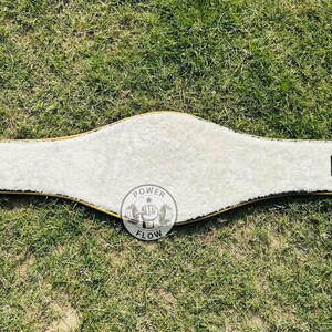 May include: A white weightlifting belt with a gold trim and a soft, plush interior. The belt has a curved design and features a circular logo with the words "POWER FLOW" and a weightlifting graphic.