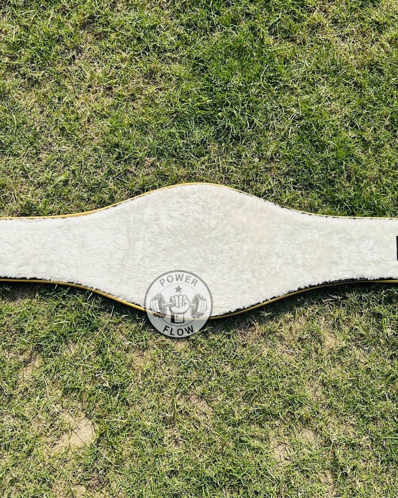 May include: A white weightlifting belt with a gold trim and a soft, plush interior. The belt has a curved design and features a circular logo with the words "POWER FLOW" and a weightlifting graphic.