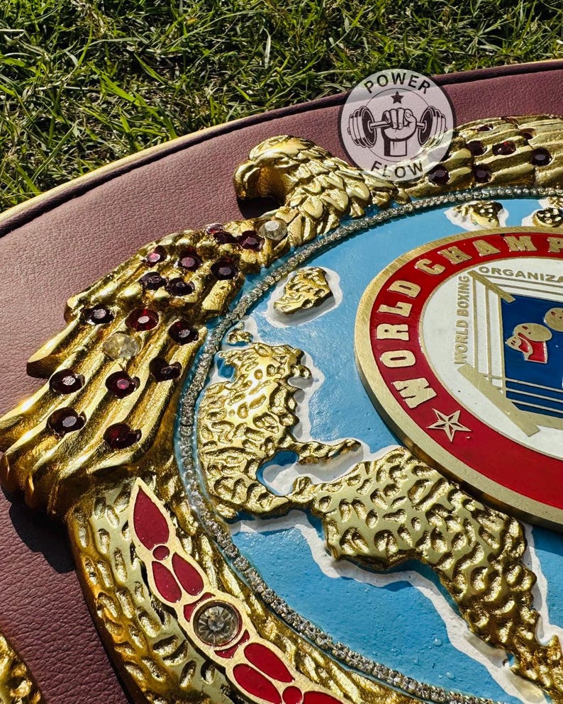 May include: A close-up of a gold and red boxing championship belt. The belt features gold eagle and leopard designs, red jewels, and the words "World Boxing Organization". The belt is on a brown surface.