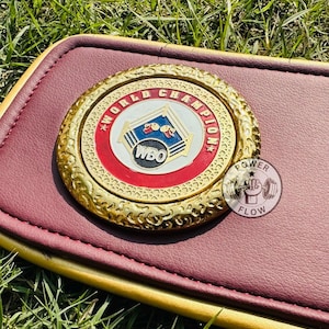 May include: A burgundy and gold boxing championship belt with a gold medallion. The medallion features the words "WORLD CHAMPION" and the WBO logo. A small, round Power Flow logo is visible.