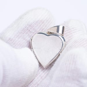 Puede incluir: Un colgante plateado en forma de corazón con un pequeño lazo en la parte superior. El colgante se sostiene con un guante blanco sobre un fondo blanco. El corazón es liso y reflectante.