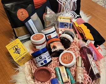 May include: A collection of Moroccan beauty and skincare products including various jars of creams, a bottle of hair oil, small containers of colored powders, a yellow box with Arabic text, a black pouch, and a pink scalp massager on a woven tray.