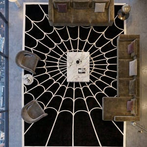 May include: Overhead view of a modern living room with a black rug featuring a white spiderweb design. The room includes a marble coffee table, two gray armchairs, and two brown sofas with decorative pillows. The room has large windows.