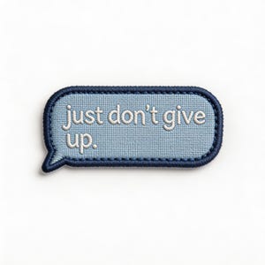 May include: A light blue embroidered patch in the shape of a speech bubble. The patch has a dark blue border and the words "just don't give up" embroidered in white. The patch is on a white background.