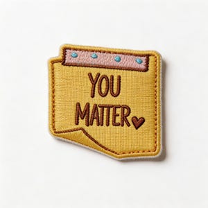 May include: Embroidered patch in a notepad shape, featuring the words "YOU MATTER" in brown lettering. The patch has a yellow background with a pink and blue border at the top and a small heart. The patch is outlined in a brown border.