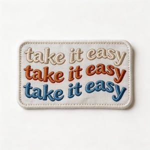 May include: Embroidered patch with the phrase "take it easy" in three rows. The top row is beige, the middle row is orange, and the bottom row is blue. The patch has a beige border and is rectangular.