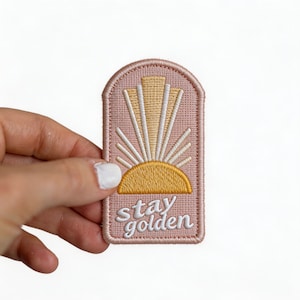 May include: Embroidered patch with a pink background and a stylized sun design in yellow and white. The patch features the words "stay golden" in white script.