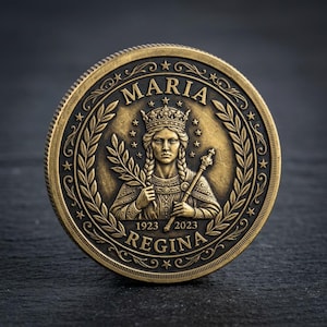 May include: A bronze-colored coin featuring a detailed relief of a woman wearing a crown and holding a scepter and a sprig. The coin is inscribed with the word "MARIA" above the image and "REGINA" below, with the years 1923 and 2023. The coin has a decorative border.