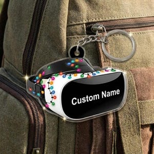 May include: A VR headset keychain with a black and white design, adorned with colorful string lights. The keychain has a silver ring and chain. The text "Custom Name" is displayed on the headset.