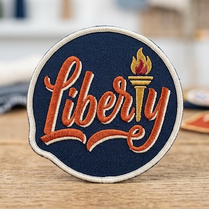 May include: Round navy blue patch with the word "Liberty" in orange script, outlined in white. A gold torch with a red flame sits above the "y". The patch has a white border and is likely an embroidered emblem.