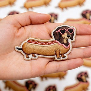 May include: Embroidered patch featuring a dachshund in a hot dog bun, wearing pink heart-shaped sunglasses. The patch has brown, tan, and pink colors. The patch is held in a hand, with other similar patches in the background.