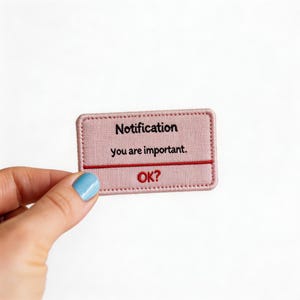 May include: A pink rectangular patch with the text "Notification you are important. OK?" in black. The patch has a red horizontal line and a stitched border. The patch is held by a hand with a blue painted nail.