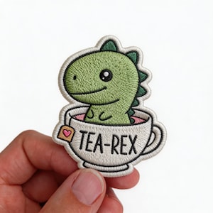 May include: Embroidered patch featuring a green dinosaur in a teacup. The dinosaur has a friendly expression and is surrounded by a white teacup with the text "TEA-REX". The patch includes a small tea bag with a heart.