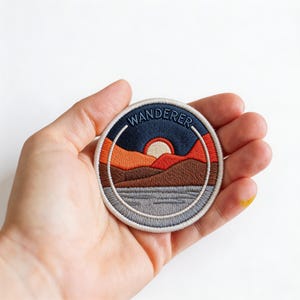 May include: Embroidered patch with a circular design featuring a sunset over mountains and a lake. The patch has a navy blue border with the word "WANDERER" in white. The sunset includes shades of orange, red, and brown. The lake is gray.